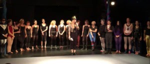 TeilnehmerInnen einer Schüler Show und eine Moderatorin mit schwarzem Zylinder Hut und Mikrofon auf einer Showbühne