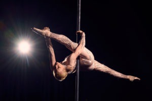 Eine Tänzerin in hautfarbenem Catsuit mit Strasssteinen macht einen Spagattrick an einem Aerial Pole