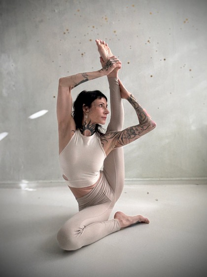 Eine Frau mit schwarzen Haaren und hellem Trainingsoutfit macht eine Stretching Pose vor einer hellen Wand.