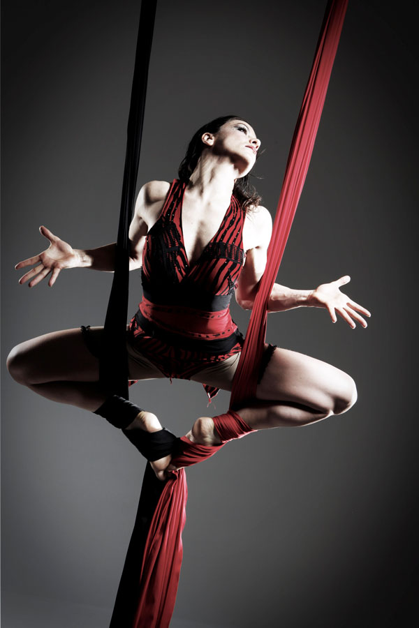 Eine Frau in schwarz-rotem Kostüm sitzt in einer Luftarkobatik-Pose in Aerial Silks vor dramatischem dunklem Hintergrund