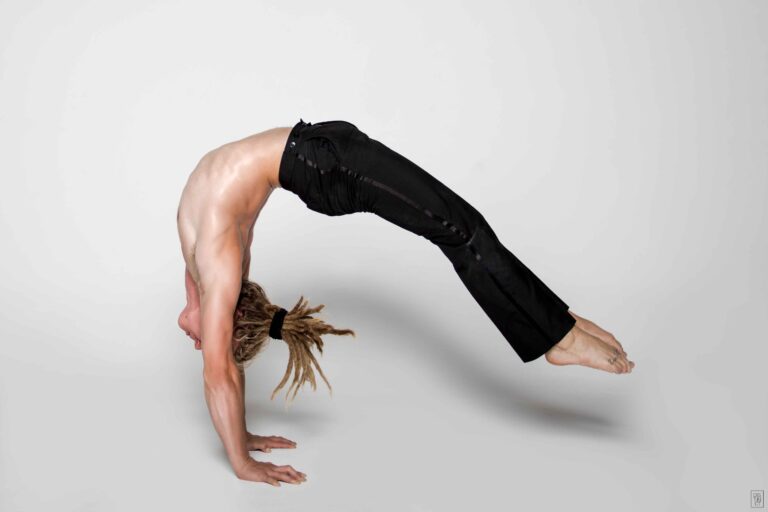 Ein Mann mit freien Oberkörper und schwarzer Hose macht einen Handstand-Trick im Backbend vor weißem Hintergrund