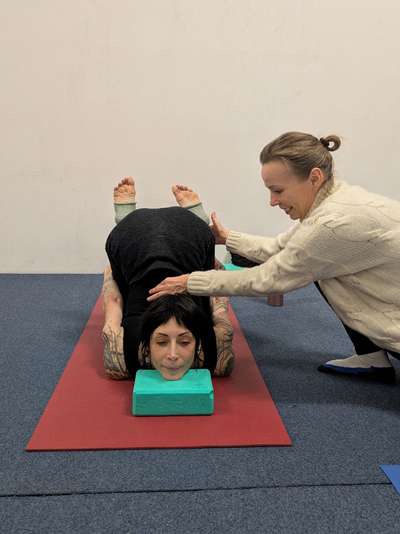 Eine blonde Trainerin in weißem Pullover korrigiert eine Schülerin im Kontorsionsunterricht, die mit dem Kinn auf einem Yogablock eine Übung für aktive Nackenflexibilität ausübt.