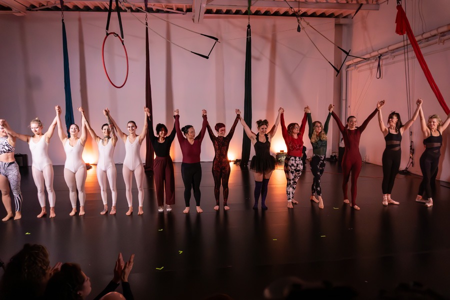 Eine Gruppe Teilnehmer:innen einer Schüler Show stehen in Kostümen Hand-in-Hand auf der Bühne eines Luftakrobatikstudios und heben die Hände zum Finale