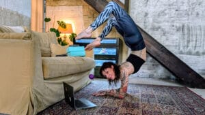 Eine Frau in Sportkleidung zeigt vor einem Laptopm am Sofa einen Unterarmstand Skorpion für einen Online Kontorsion Kurs